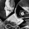 A black and white photograph showing a foot wearing silver high heels next to a diamond-encrusted clutch and sheer fabric wit