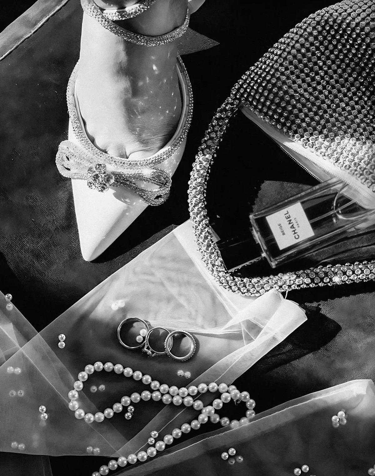 A black and white photograph showing a foot wearing silver high heels next to a diamond-encrusted clutch and sheer fabric wit
