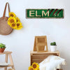 A vintage metal tin sign with the text ELM Str. in green on a white wall, decorated with sunflowers.