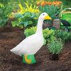 A white goose garden statue with an orange beak and green feet is placed in dark soil among green plants.