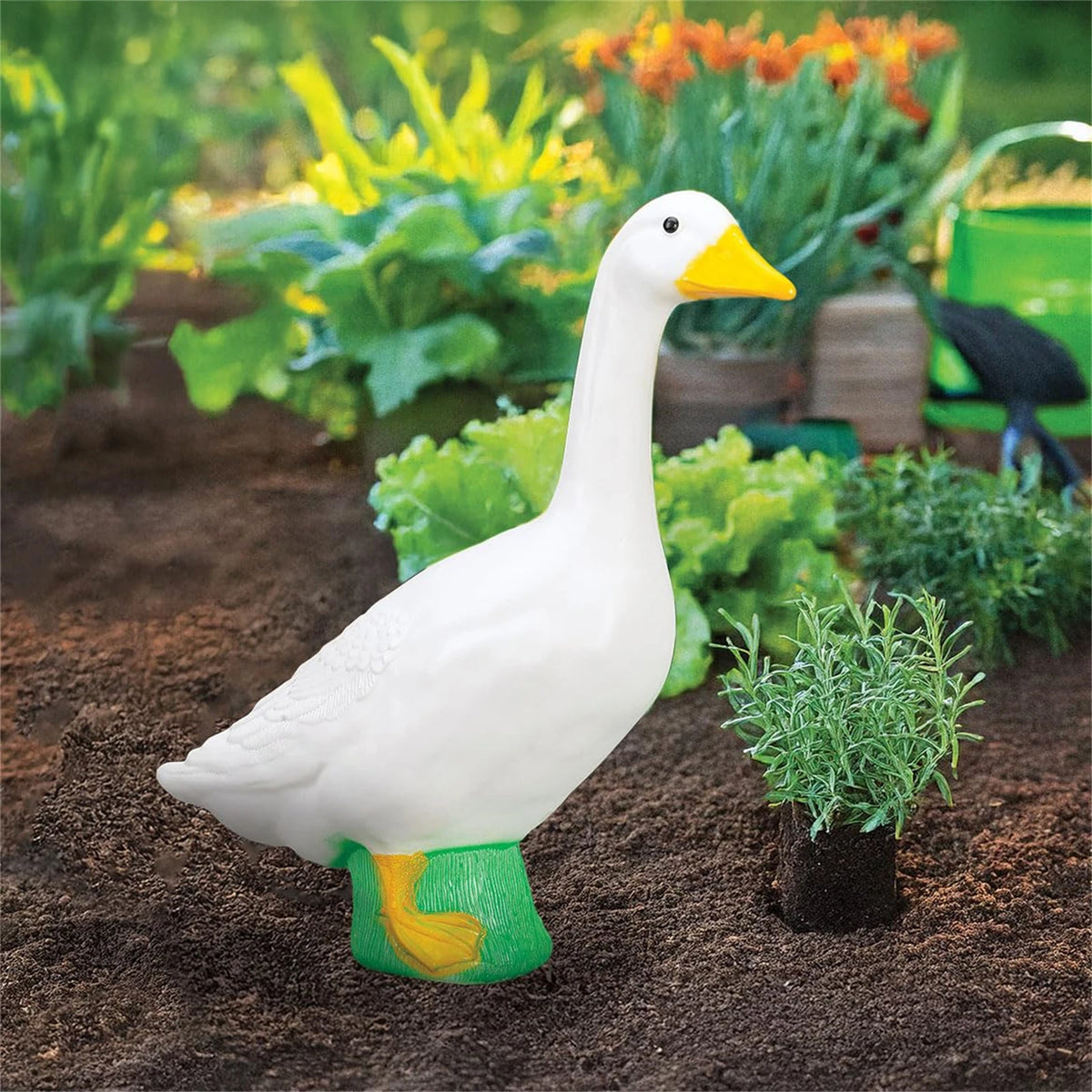 A white goose garden statue with an orange beak and green feet is placed in dark soil among green plants.