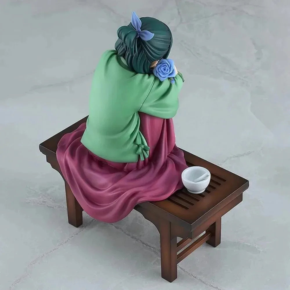 A collectible PVC figure of Maomao from The Apothecary Diaries, depicted with green hair and a pink robe, sitting on a wooden