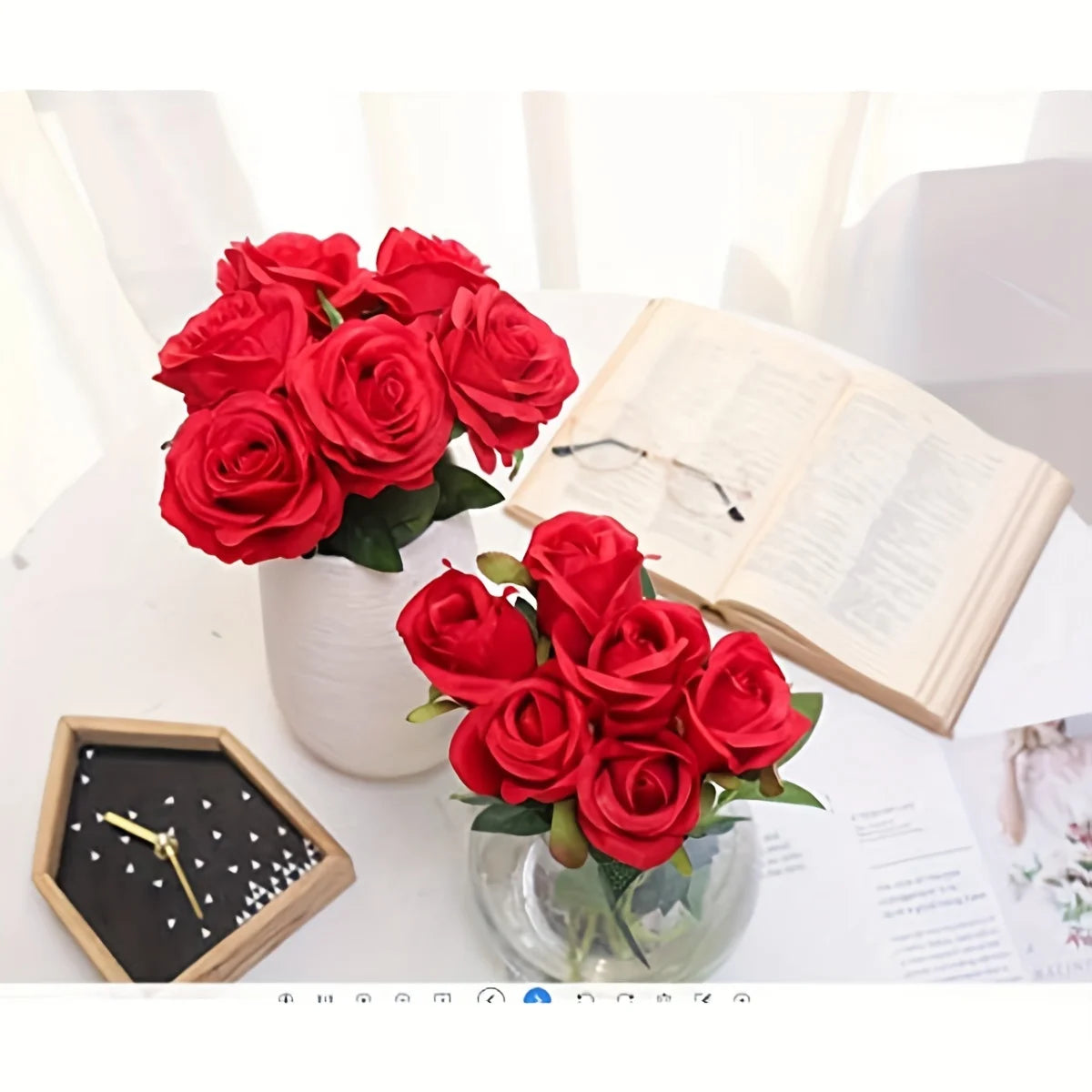 Un ramo de rosas artificiales rojas de seda en un jarrón de vidrio junto a un libro abierto y un reloj de madera.