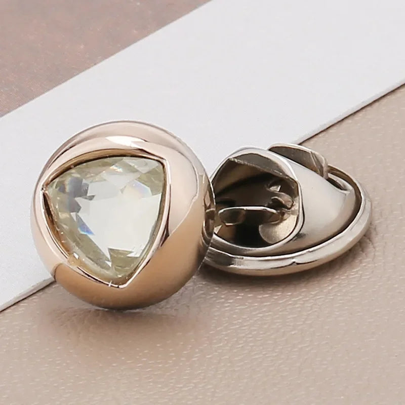 A close-up of a rose gold brooch button featuring a clear, faceted gemstone set in a diamond shape.