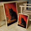 A framed poster depicting a black cat smoking on a wooden surface, displayed next to a light wood cabinet.