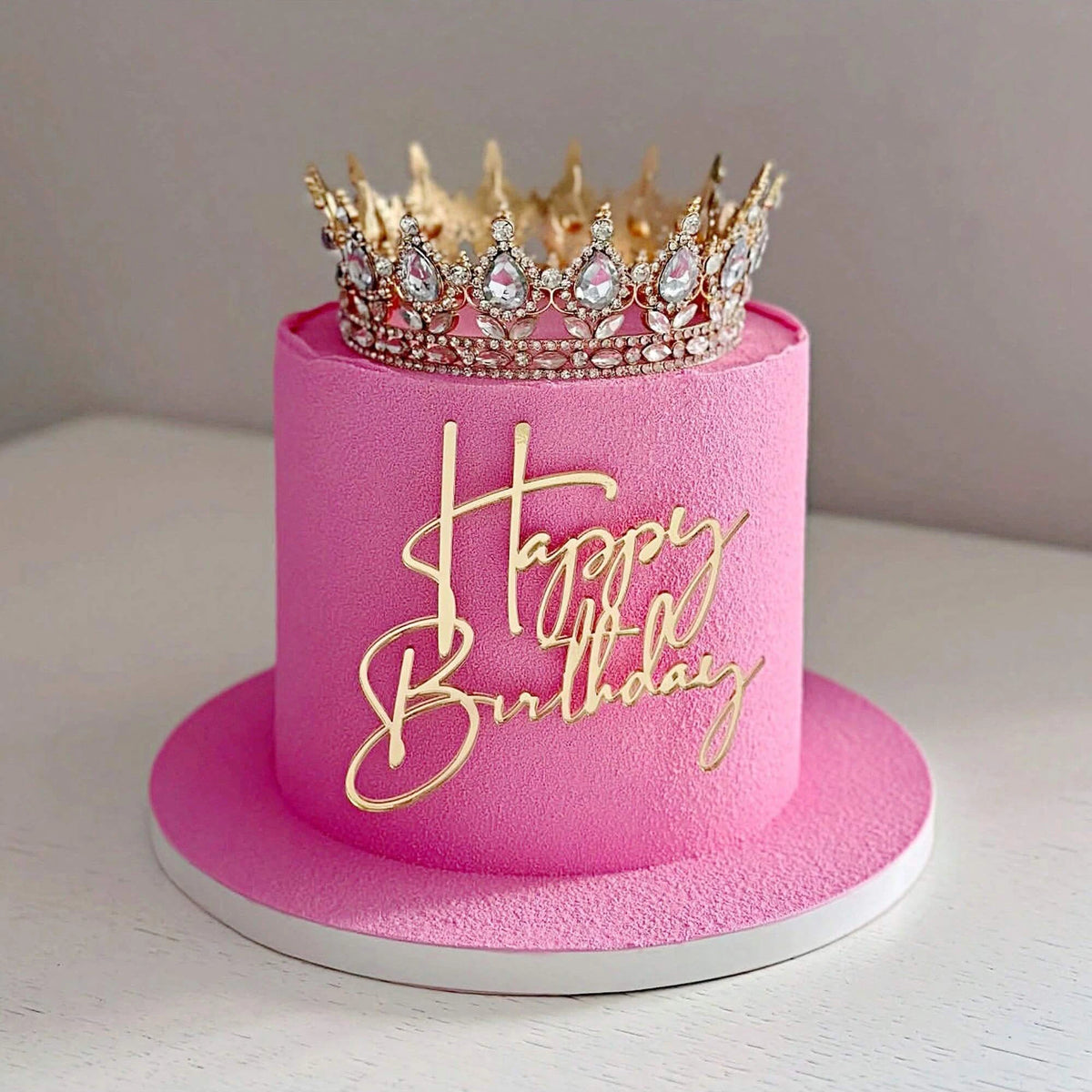 A pink birthday cake with a gold glitter topper featuring the words Happy Birthday