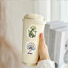 A hand holds a beige insulated tumbler with a cute doodle sticker featuring a tree and a figure.
