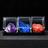 Three rough crystal specimens in purple, blue, and red are displayed in clear acrylic boxes.