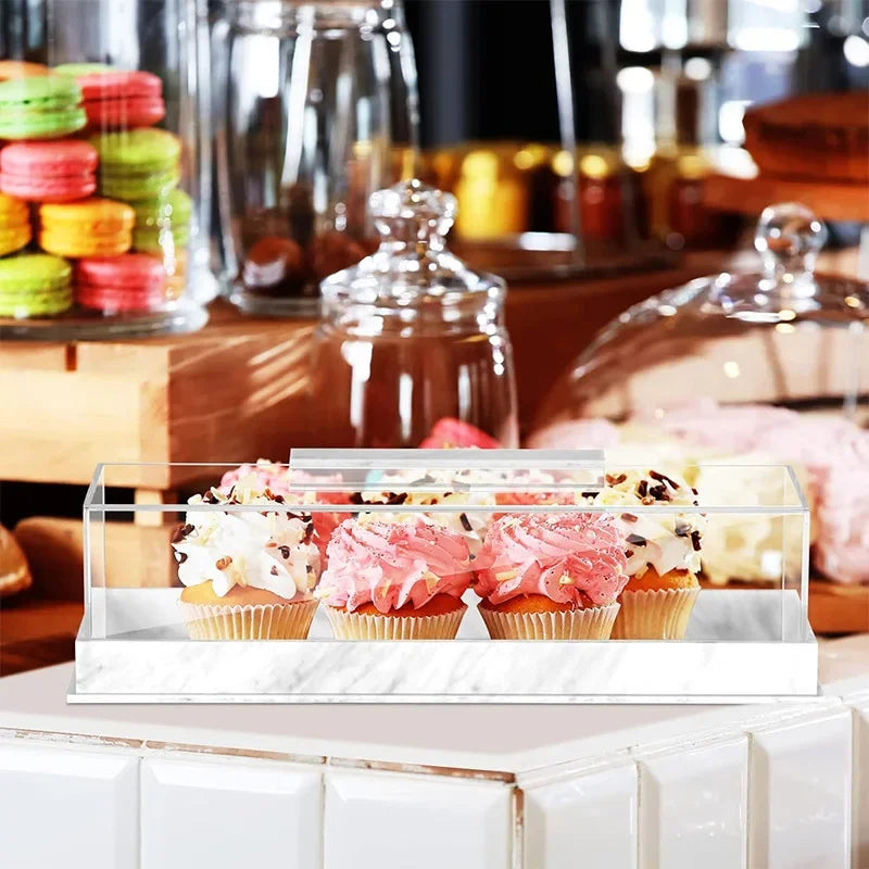 Clear acrylic cake stand with a lid displaying pink and yellow cupcakes on a marble surface.