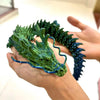 A hand holds a green and blue articulated 3D printed dragon figurine with textured scales.
