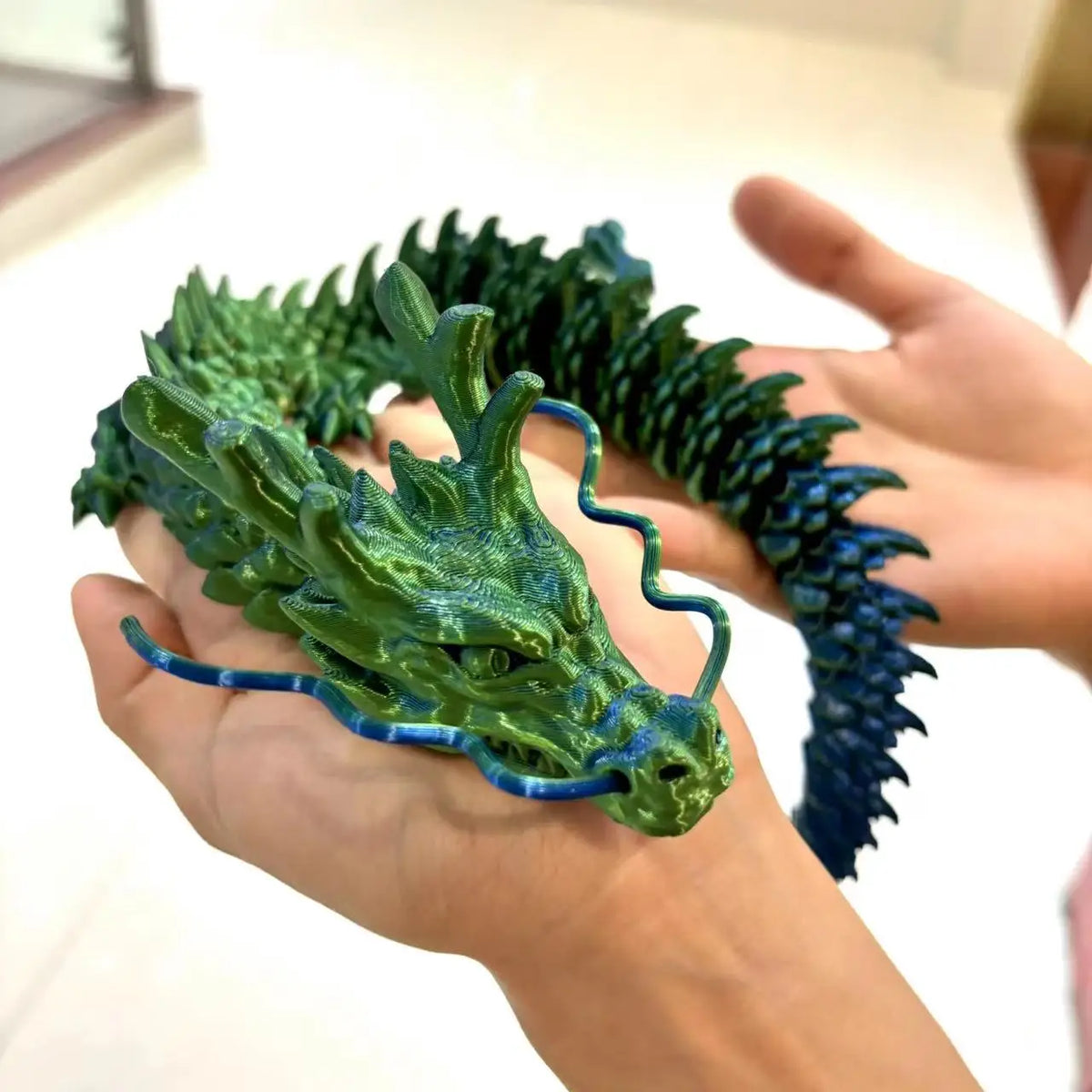 A hand holds a green and blue articulated 3D printed dragon figurine with textured scales.