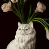 A white ceramic cat sculpture with long fur sits among pink tulips.