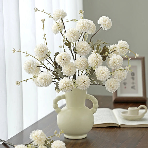 A bouquet of white silk chrysanthemum artificial flowers in a light ceramic vase on a wooden table.