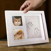 A white photo frame displays a cat's face and a clay paw print mold, held by a hand.