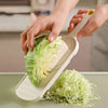 A hand is using a manual cabbage shredder to process a head of green cabbage into shreds.