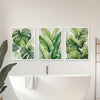 A set of three watercolor leaf art prints featuring lush green tropical foliage displayed on a white wall above a bathtub.