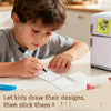 A young boy is drawing designs on paper with a pen, preparing to make DIY acrylic fridge magnets.