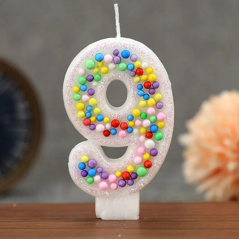 A white birthday candle shaped like the number 9 decorated with colorful glitter candy beads