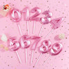 Pink birthday candles shaped like numbers 0 through 9 arranged on a pink background.