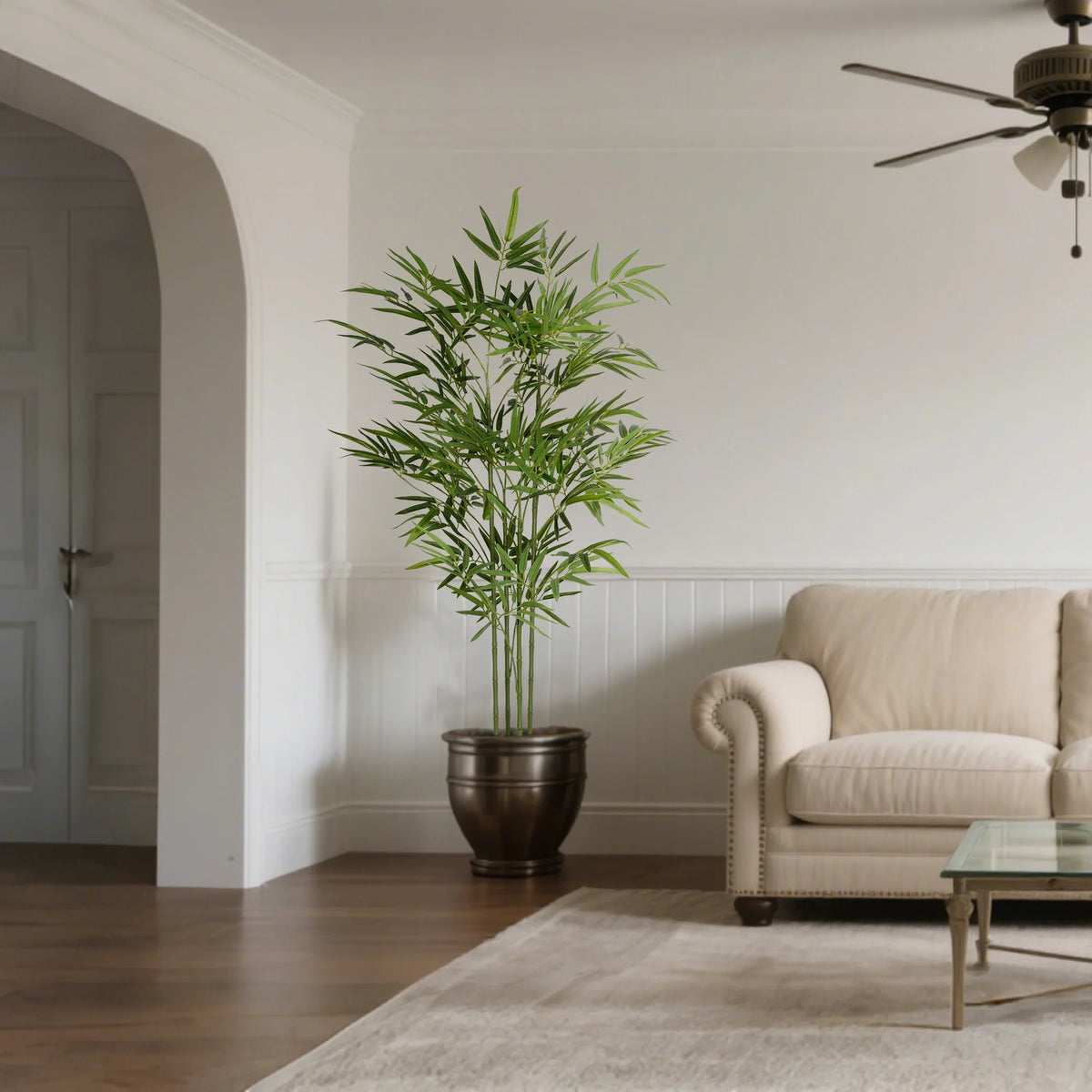 An artificial bamboo tree in a dark pot stands on the floor next to a light-colored sofa and glass table.