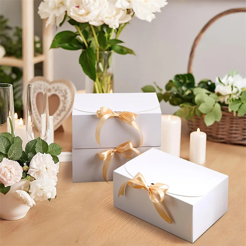 White kraft paper gift boxes tied with gold ribbon resting on a wooden table with flowers and greenery.