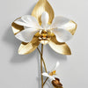 Flor de orquídea blanca prensada con hoja de oro en un tallo delgado contra una pared gris claro.