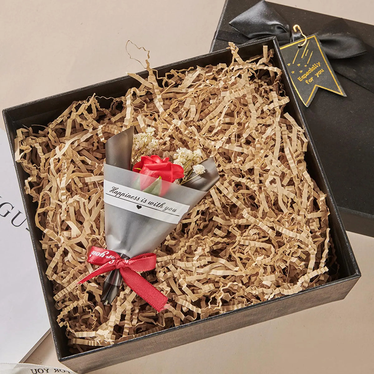 A gift box filled with crinkle cut paper shred filler containing a small arrangement of red roses and a card