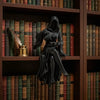 A black resin statue of a female grim reaper wearing a hooded cloak and holding a scythe, displayed on a wooden bookshelf.