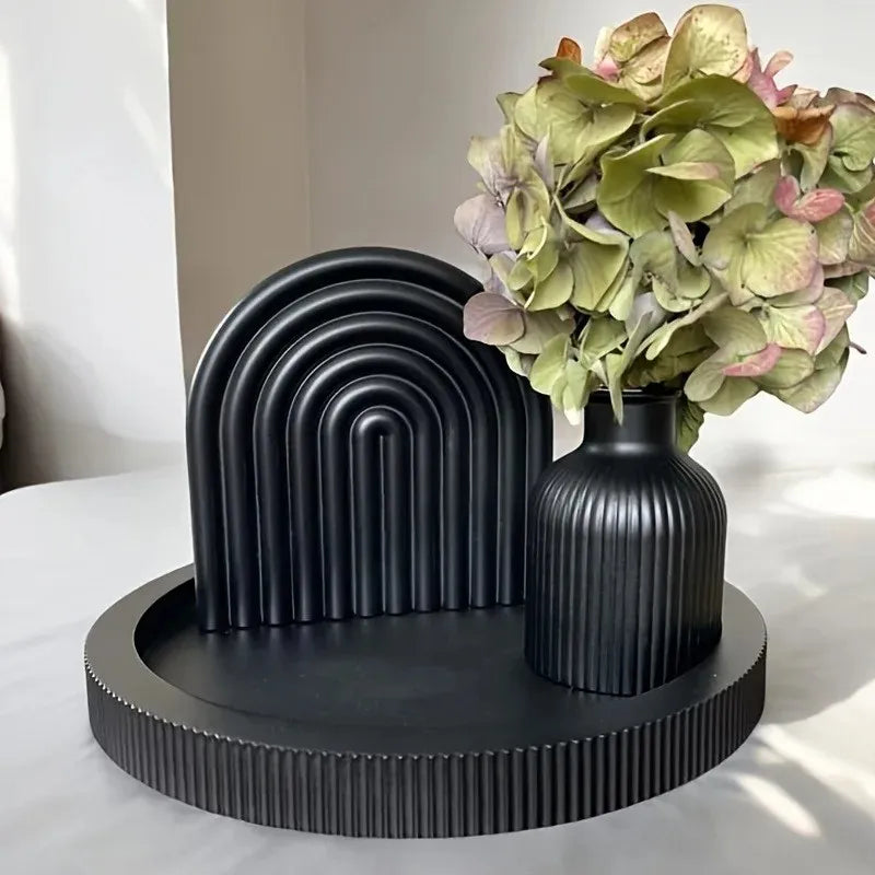 A black silicone mold for vases and trays is displayed on a round black wooden base with a bouquet of green and pink foliage.