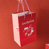 A red gift box with pink accents and a surprise tag is held by a hand against an orange background.