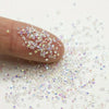 A finger is sprinkling iridescent nail rhinestones onto a white surface.
