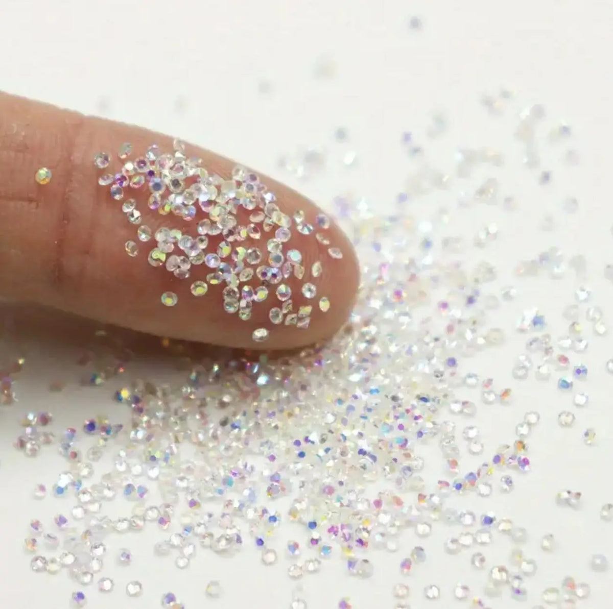 A finger is sprinkling iridescent nail rhinestones onto a white surface.