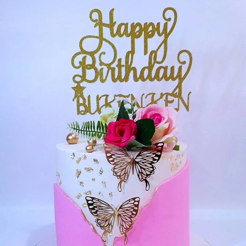 A white cake decorated with gold butterfly designs and a gold script topper that reads Happy Birthday