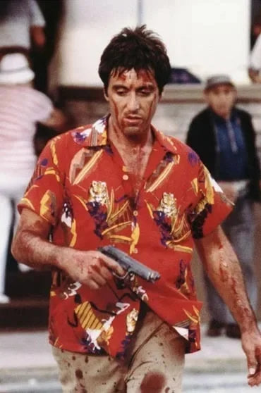 Un homme en chemise rouge à motifs tient une arme de poing, probablement du film Scarface, dans une scène dramatique.