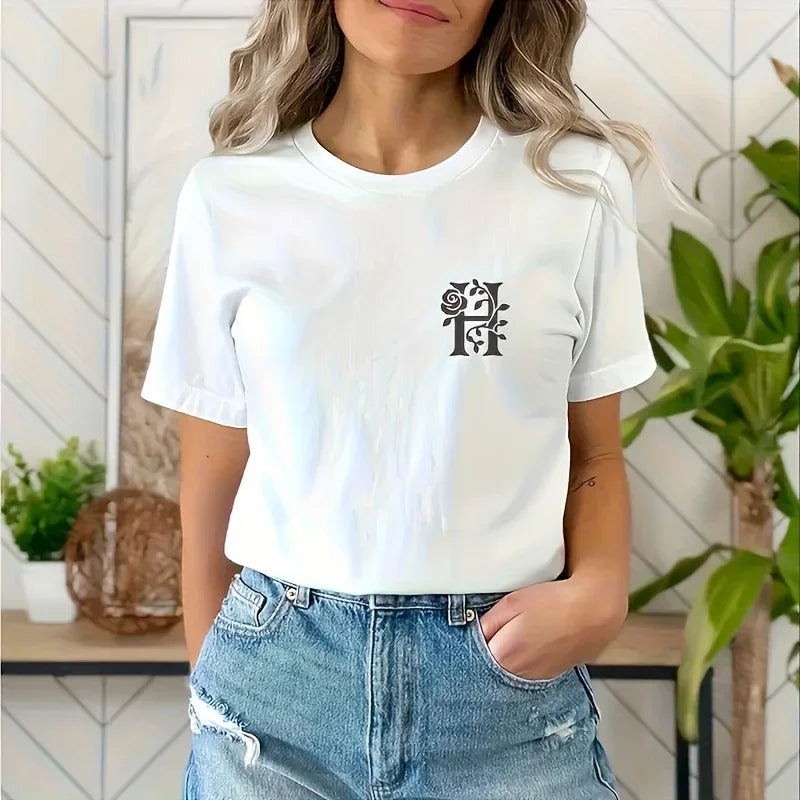 A woman models a white t-shirt featuring a black floral alphabet stencil design, paired with blue jeans.
