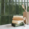 A display of natural soap bars and wooden toothbrushes against a dark green tiled backsplash.