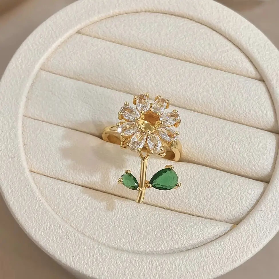 A gold cocktail ring featuring a floral design with clear stones and two green leaf accents.
