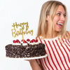 A blonde woman holds a black cake with gold birthday topper letters against a white background.