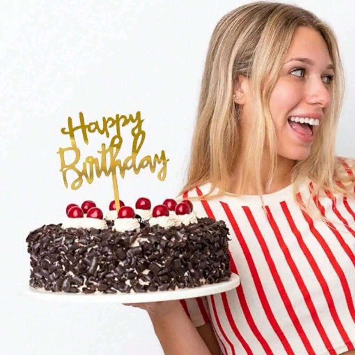 A blonde woman holds a black cake with gold birthday topper letters against a white background.