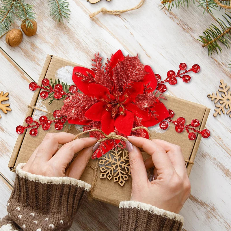 Hands are placing red glittered artificial Christmas flowers with clips onto a brown gift box decorated with pine branches an