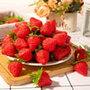 A plate of bright red artificial strawberries arranged on a wooden board with greenery.
