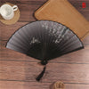 A dark gray lace folding hand fan with delicate white floral embroidery rests on a wooden surface next to a cup and a straw m