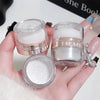 A hand holds various glass jars of white and glitter body highlight powder products.
