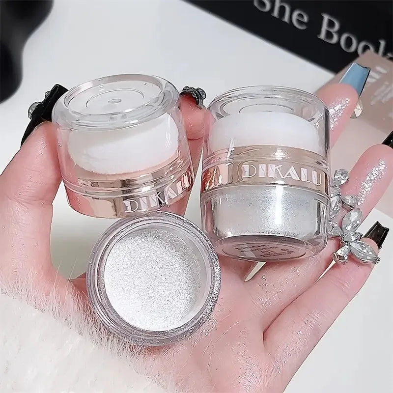 A hand holds various glass jars of white and glitter body highlight powder products.