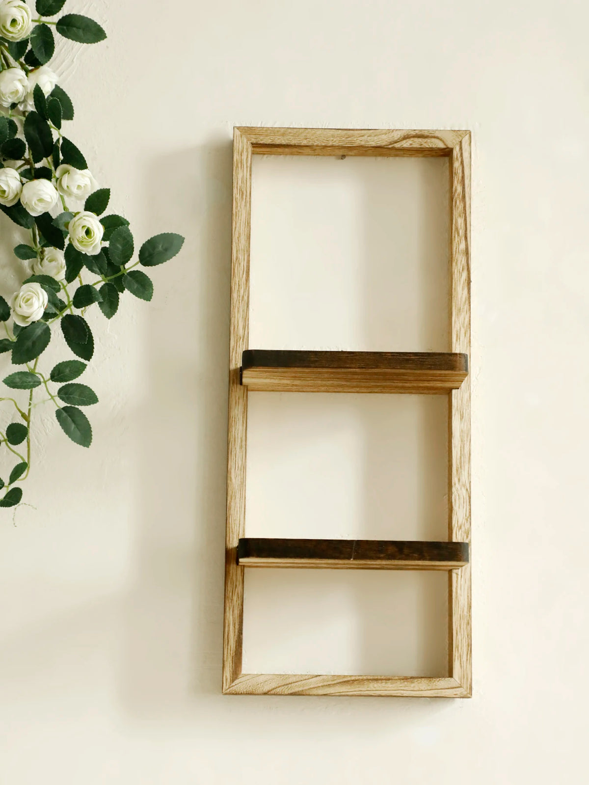 A light wood floating wall shelf with two tiers mounted on a white wall, decorated with green leaves and white roses.