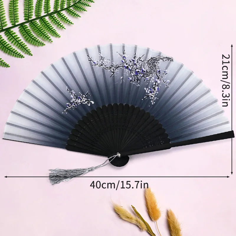 Elegant black and silver bamboo folding hand fan with a tassel displayed against a pink background.