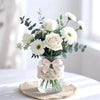A bouquet of white roses and greenery arranged in a clear glass vase on a woven mat.