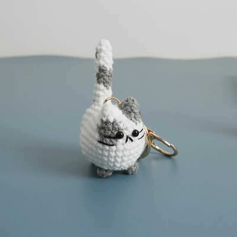 A handcrafted crochet cat keychain pendant with gray and white fur resting on a smooth blue-gray surface.