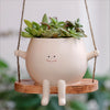 A small, light pink resin succulent planter with a smiling face is suspended by a twine hanger over a wooden swing base.
