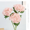 A bouquet of pink artificial hydrangeas with green leaves displayed against a light gray background.